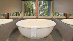 Une grande baignoire sur pieds se trouve dans une salle de bain équipée de deux lavabos et d’une fenêtre donnant sur la forêt.