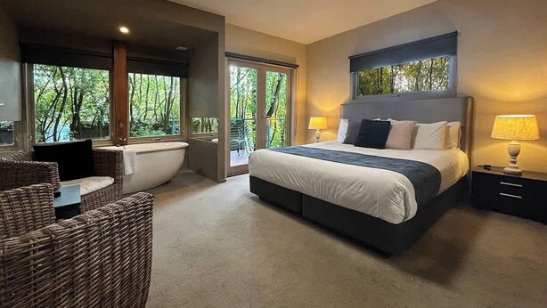 Une chambre moderne avec un grand lit, un coin salon avec des meubles en osier et une vue sur la verdure par les fenêtres.