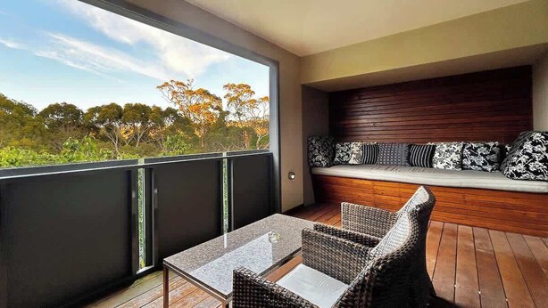 A balcony with a glass door, wicker furniture, and a built-in bench with patterned cushions. - The Retreat at Villa 7 (Hepburn Springs)