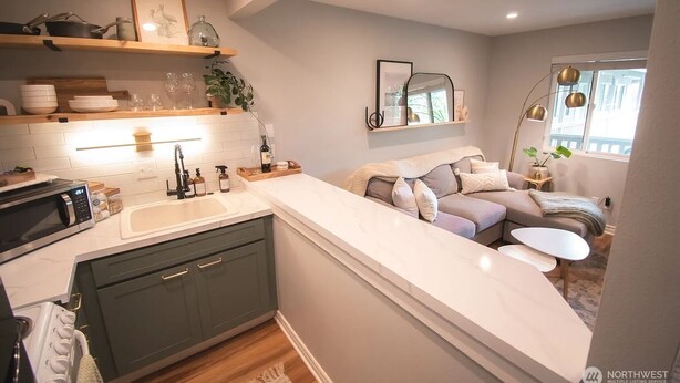 Una cocina moderna con encimera blanca, armarios oscuros y horno empotrado. Hay una zona de comedor con sofá y una mesa pequeña.