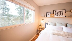 Un dormitorio con un ventanal grande, una cama con ropa de cama blanca, una mesita de noche con una lámpara y una obra de arte en la pared.
