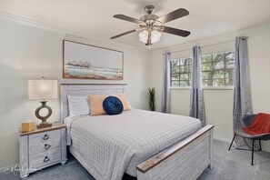 A bedroom with a bed, a nightstand, a lamp, a ceiling fan, a window with curtains, and a chair.
