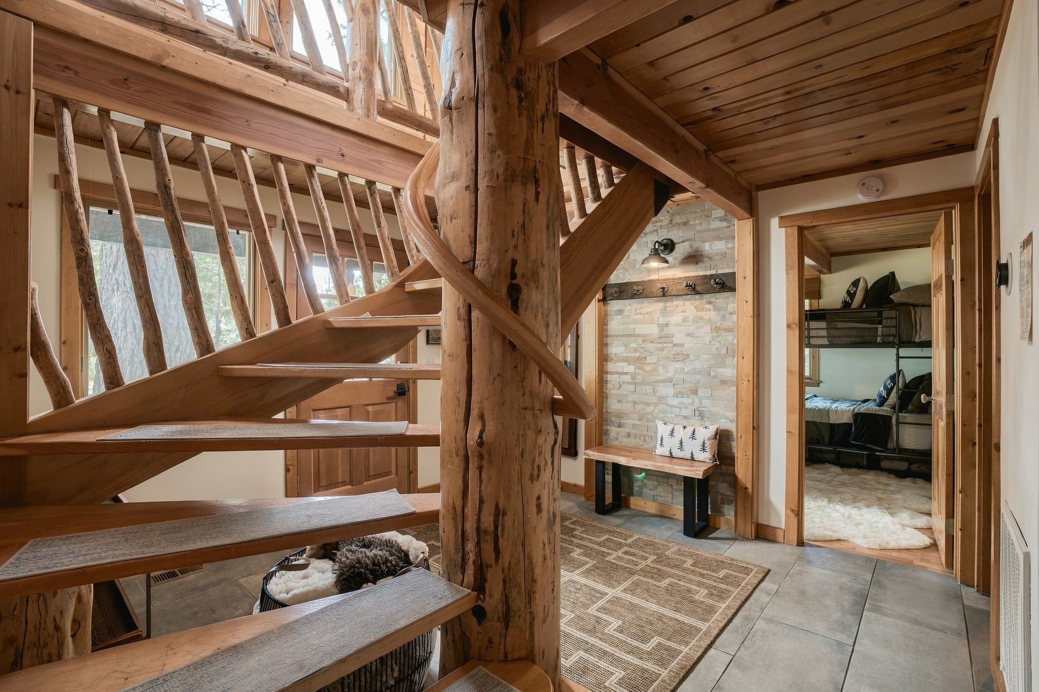 A wooden spiral staircase in a rustic interior with a stone wall and a bench.