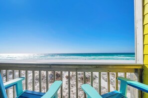Una terraza de madera con sillas turquesa con vista a una playa de aguas azules y arena.