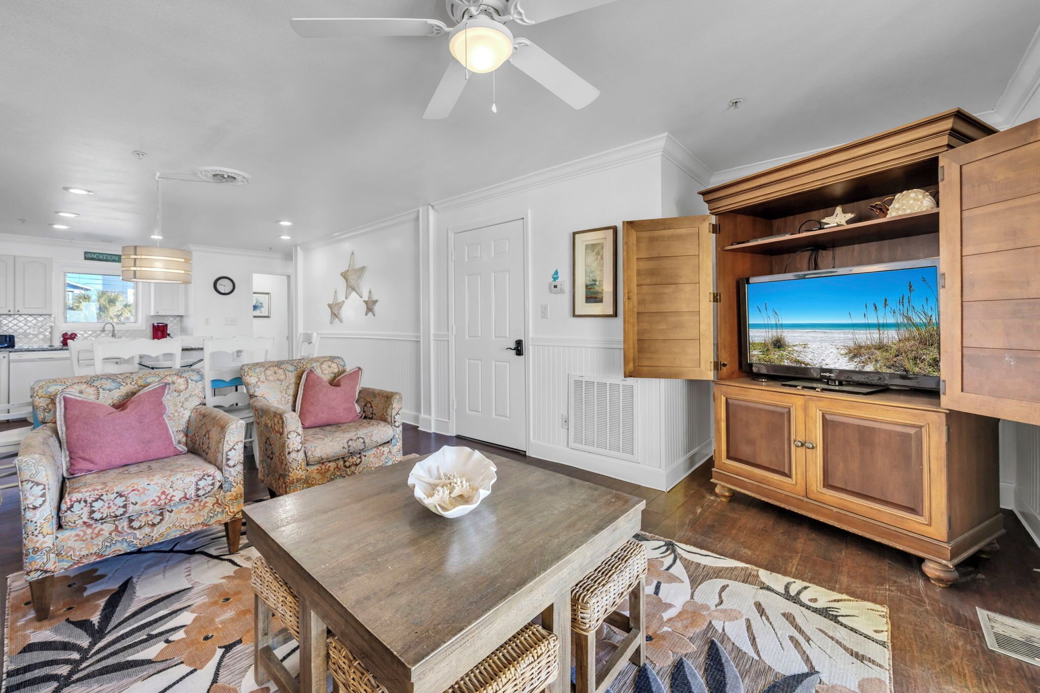A living room with a wooden coffee table, two patterned armchairs, a television on a wooden cabinet, and a large window with a view of the ocean.