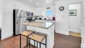 A modern kitchen with white cabinetry, stainless steel appliances, a granite countertop island, and a wooden stool. - Gulf Front- Directly On The Beach- Sunchase 2A (Miramar Beach)