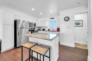 A modern kitchen with white cabinetry, stainless steel appliances, a granite countertop island, and a wooden stool. - Gulf Front- Directly On The Beach- Sunchase 2A (Miramar Beach)