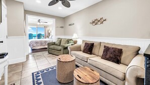 Een ruime woonkamer met een beige bank, twee fauteuils, een plafondventilator en uitzicht op de oceaan door de open deur.