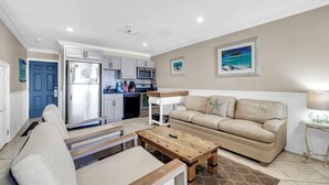 A modern living room with a beige sofa, a wooden coffee table, and a kitchen in the background. - Honeymoon Studio* Directly On Sand* Sunchase 3B (Miramar Beach)