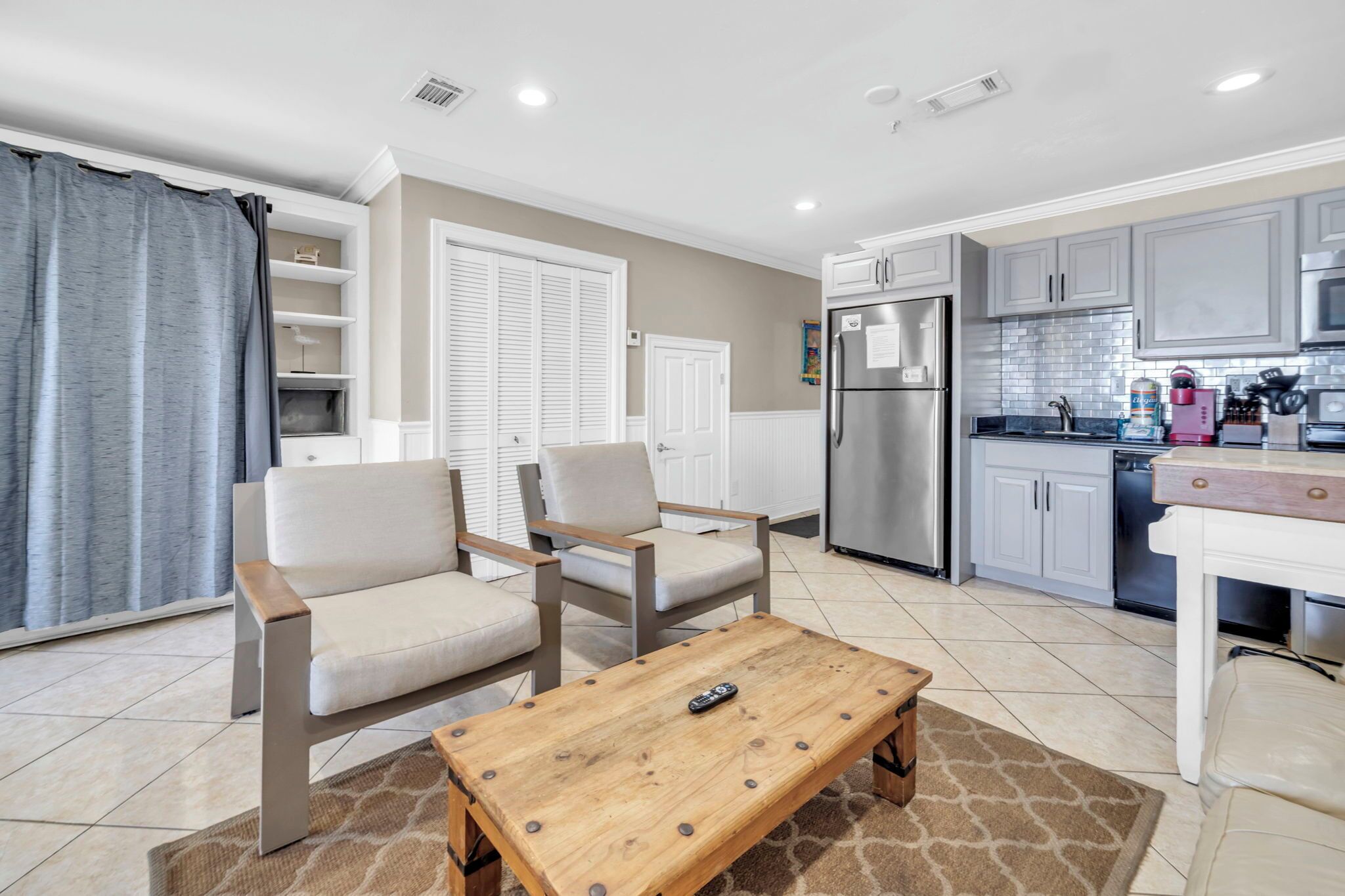 A modern kitchen with grey cabinets, a stainless steel refrigerator, and a coffee maker. There is a wooden coffee table with a remote control on it.