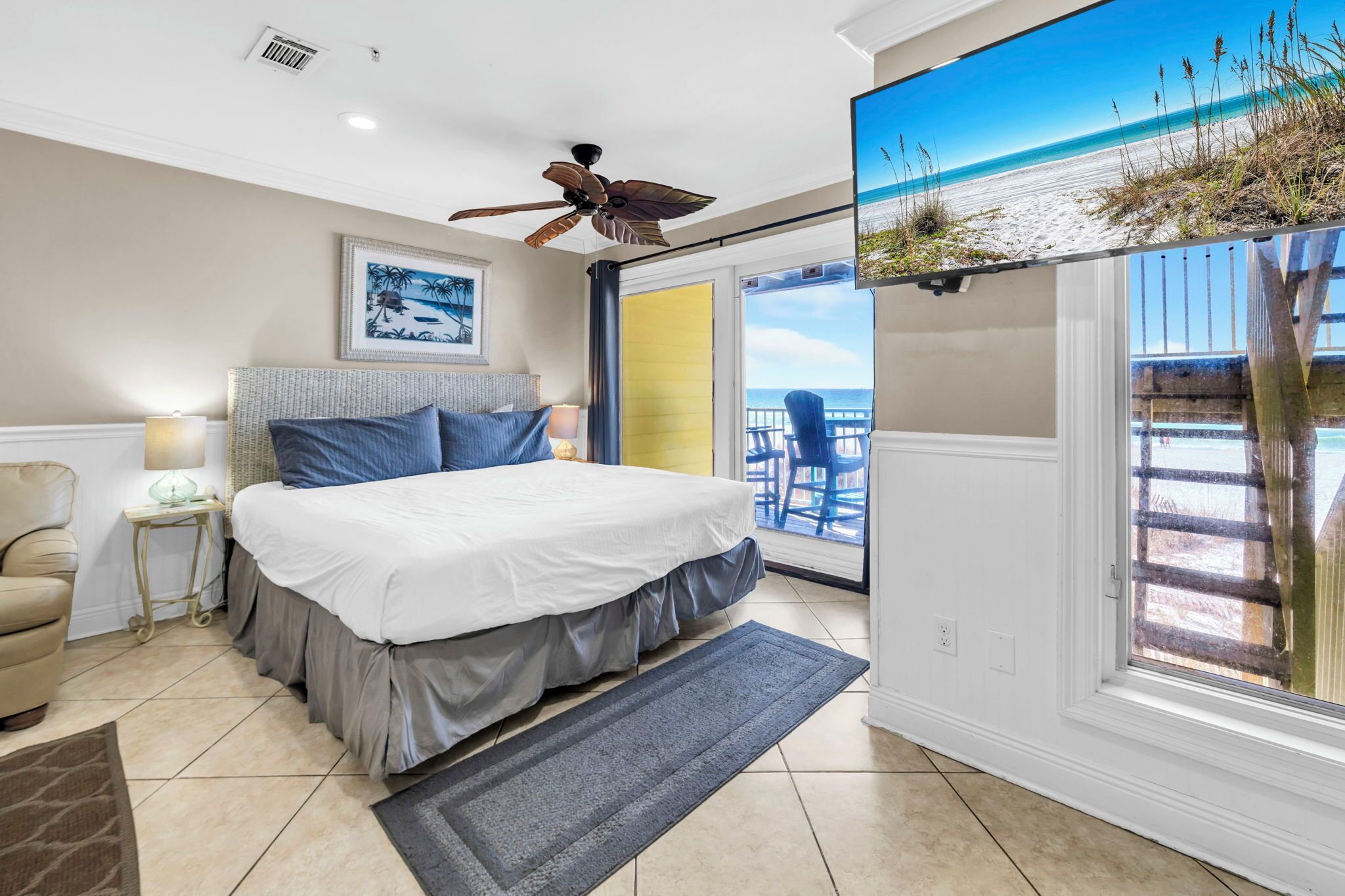 A bedroom with a large bed, a ceiling fan, a window with a view of the beach, and a chair.