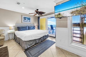 Une chambre avec un grand lit, un ventilateur de plafond, une fenêtre donnant sur la plage et un fauteuil.