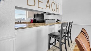 A kitchen with a white island, bar stools, and a 'BEACH' sign above the counter. - Sunchase 7A - Directly On The Beach! Sleeps 8-Walk to Restaurants, AMAZING VIEWS! (Miramar Beach)