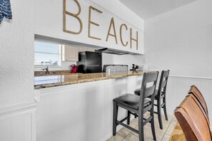 A kitchen with a white island, bar stools, and a 'BEACH' sign above the counter. - Sunchase 7A - Directly On The Beach! Sleeps 8-Walk to Restaurants, AMAZING VIEWS! (Miramar Beach)