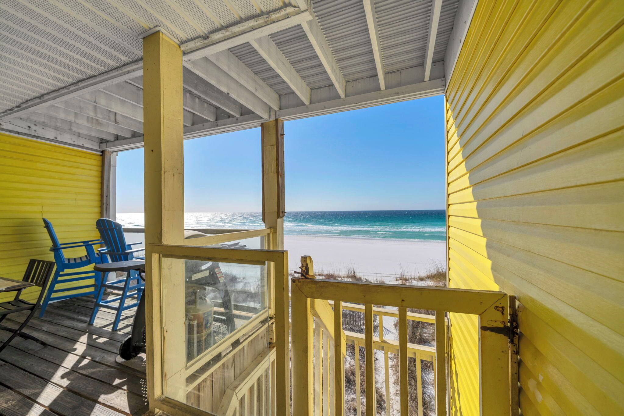 A covered outdoor area with a view of the beach, featuring yellow walls, blue chairs, and a wooden deck.