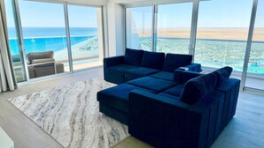 Ett modernt vardagsrum med en hörnsoffa, ett stort fönster med utsikt över stranden och havet samt en skjutdörr i glas som leder ut till ett utomhusområde.