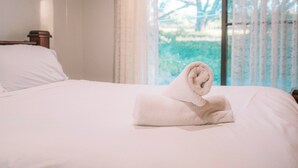 Un lit bien fait, avec une serviette enroulée posée au centre, une tête de lit en bois et une fenêtre ornée de rideaux légers.