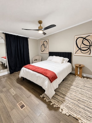 Un dormitorio con cama, ventilador de techo, silla, alfombra y obras de arte en las paredes.