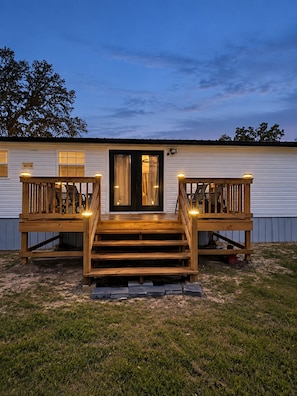 A two-story wooden deck with a staircase leading to a dark-colored door, surrounded by a grassy area.