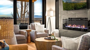 Un salon moderne doté d’une cheminée, de grandes fenêtres offrant une vue sur les arbres et les montagnes, et d’un coin salon confortable avec des canapés à carreaux et une table basse ronde en bois.