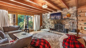 A cozy cabin interior with a stone fireplace, wooden beams, and a large window overlooking a forest. - Idyllwild Retreat - Fire Pit, Views, Game Room (Idyllwild-Pine Cove)