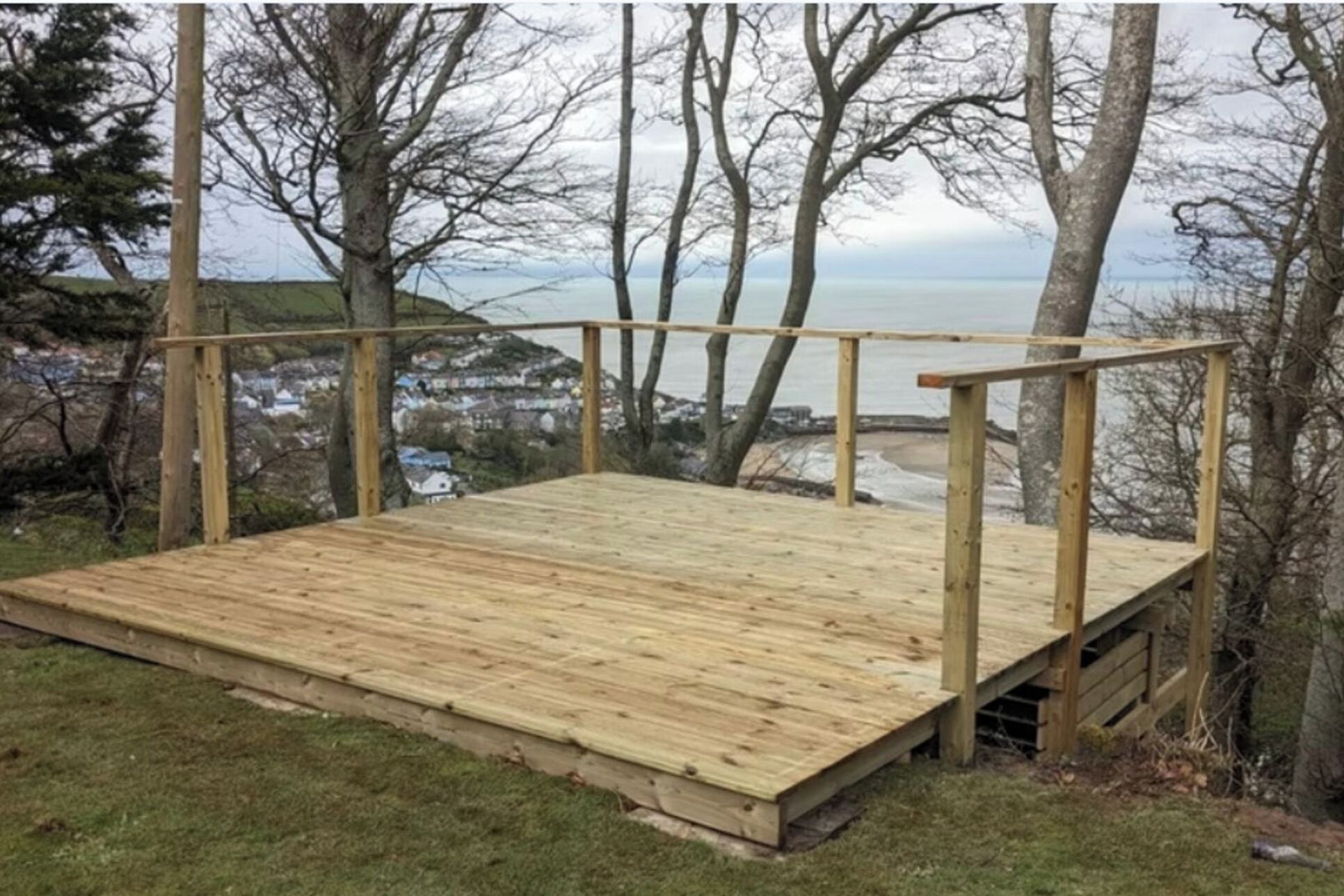 A wooden deck with railings, surrounded by trees and overlooking a coastal area.