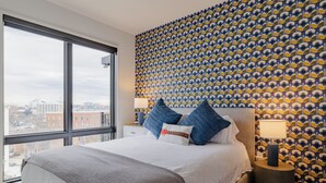 Quarto de hotel com uma grande janela, papel de parede estampado, cama com edredom cinza e branco, e mesa de cabeceira com abajur.