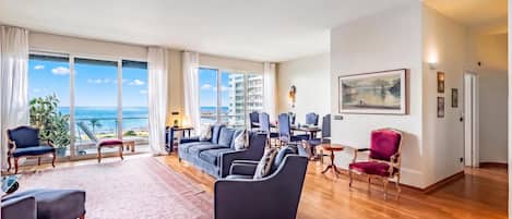 A spacious living room with a large window offering a view of the sea, a dining area with chairs and a table, and a red armchair.