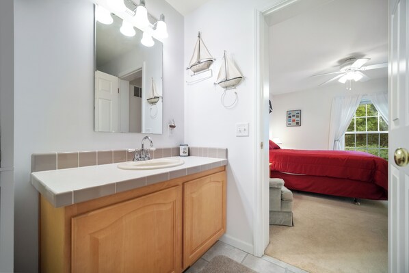 Baño con lavamanos, espejo y armarios de madera. En la habitación contigua hay una cama roja.