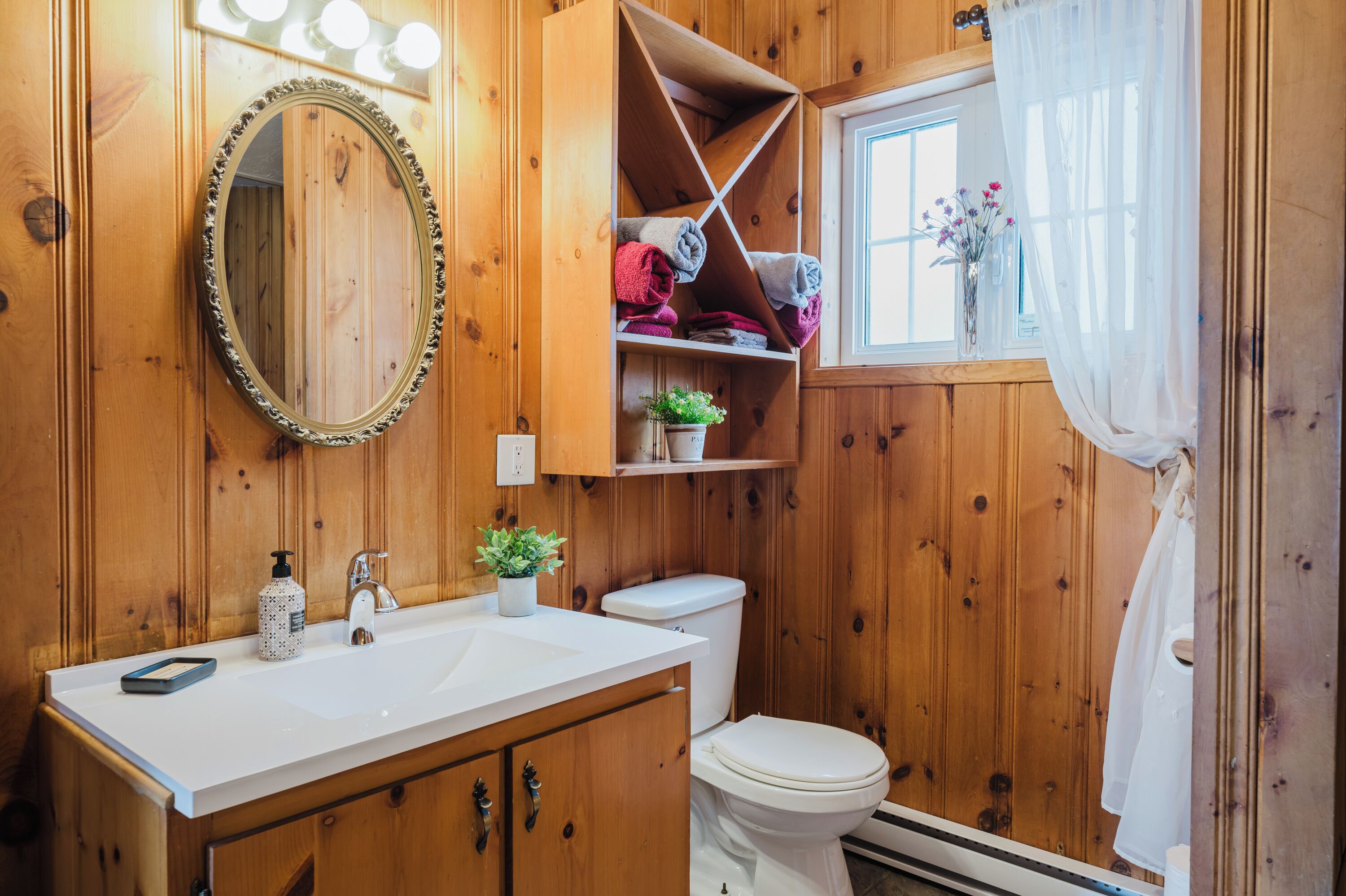 A wooden bathroom with a white sink, mirror, and toilet. There are shelves with towels and a potted plant.