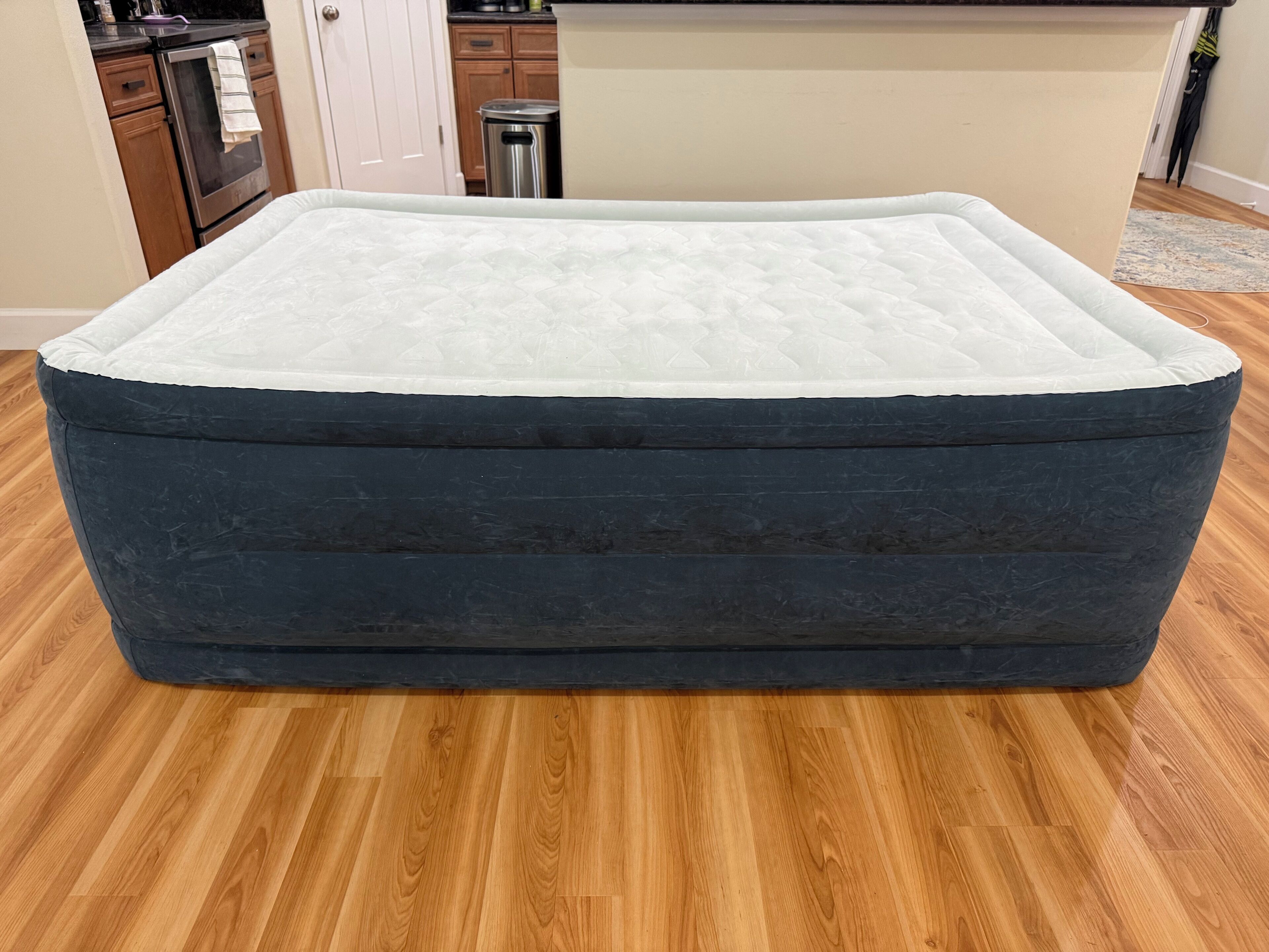 A large, dark-colored, rectangular mattress with a white top layer, placed on a wooden floor in a room with wooden cabinets and a stainless steel oven in the background.