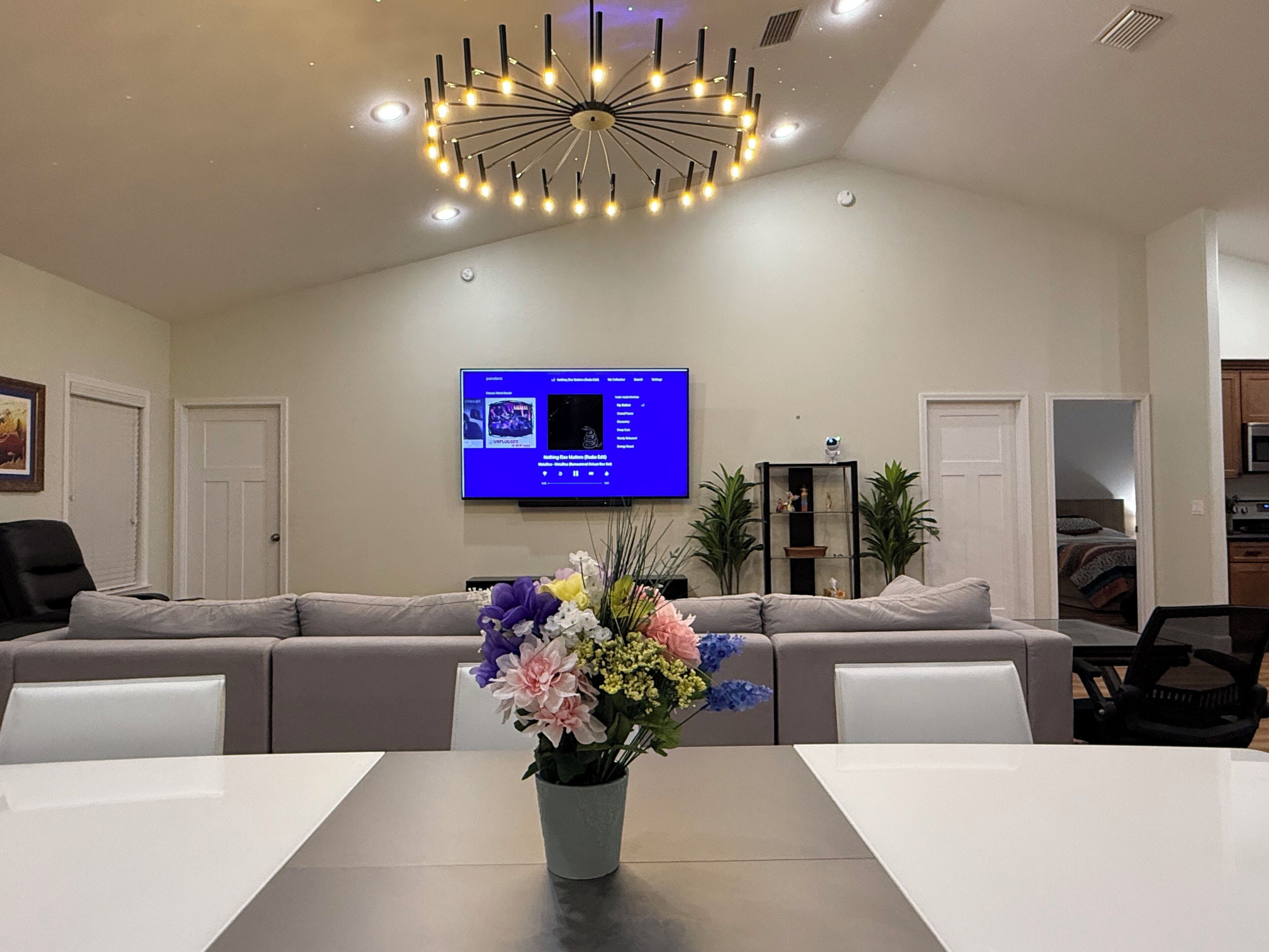 Une salle de conférence avec une longue table, un lustre moderne, un téléviseur accroché au mur et un canapé au fond de la pièce.