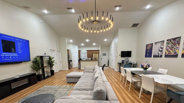 Uma sala moderna com um sofá seccional, uma TV de tela plana, uma área de jantar com mesa e cadeiras, e uma cozinha ao fundo.