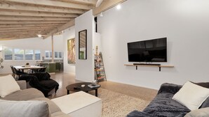 Ein modernes Wohnzimmer mit Fernseher, Sofas und einem Couchtisch. Im Hintergrund ist eine Küche mit Essbereich zu sehen.