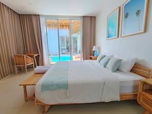 A hotel room with a large bed, a wooden nightstand, a wicker chair, and a view of a swimming pool. - Villa Chioni Uluwatu (Ungasan)