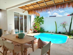 Zona de piscina con mesa y sillas, techo de paja y diversas plantas.