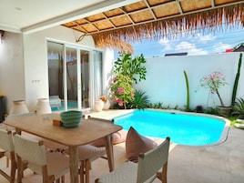 Zona de piscina con mesa y sillas, techo de paja y diversas plantas.