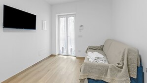 A modern, minimalistic living room with a flat-screen TV mounted on the wall, a sliding glass door leading to a balcony, and a blue sofa with a blanket. - Merlo A -  Center & Private Parking, Ceriale, Italy (Ceriale)