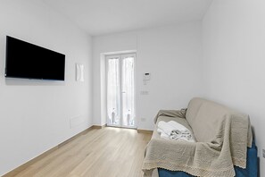 Ein modernes, minimalistisches Wohnzimmer mit einem an der Wand befestigten Flachbildfernseher, einer Schiebetür zu einem Balkon und einem blauen Sofa mit Decke.