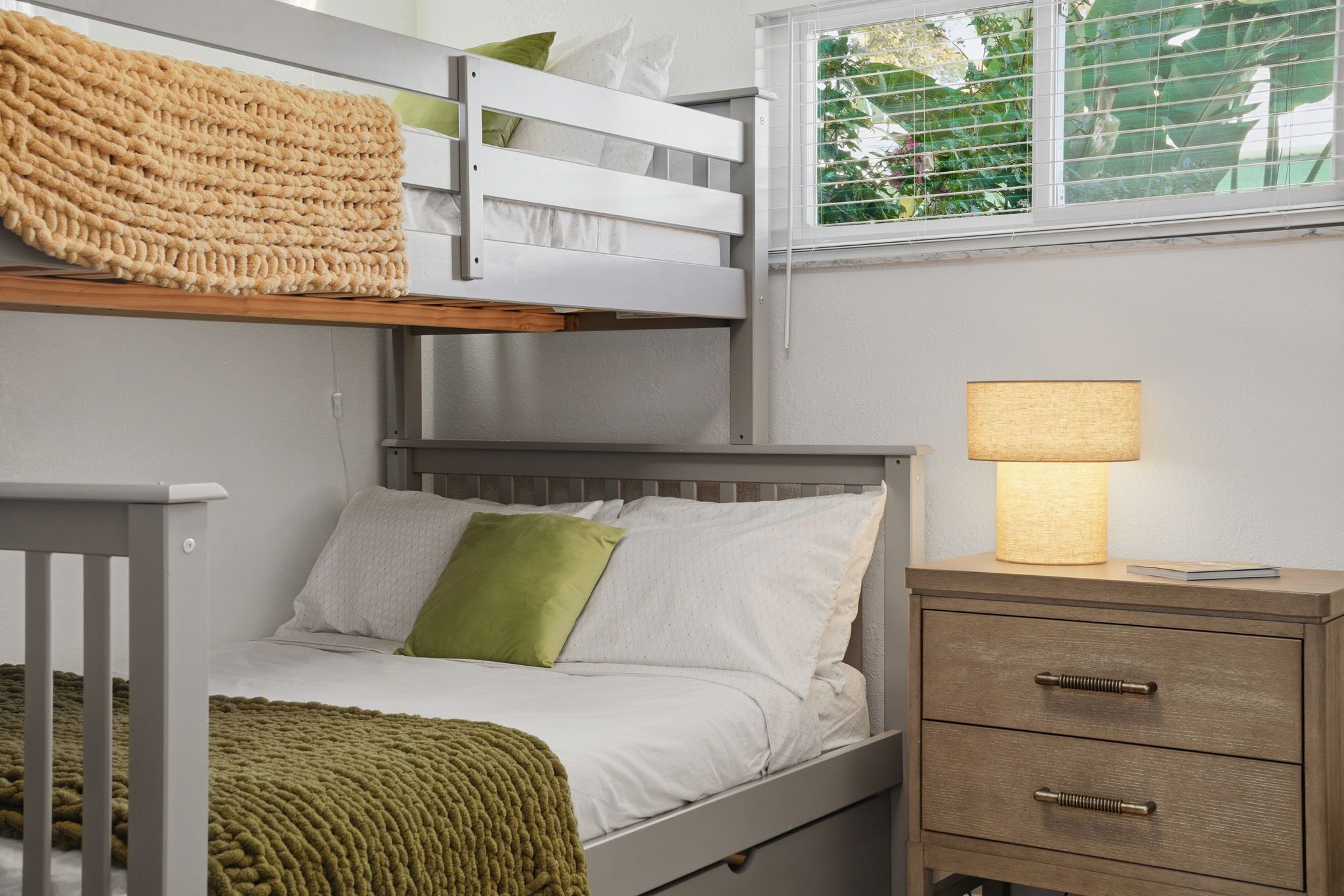 Una cama individual superpuesta con una manta verde, una lámpara de noche y una mesita de noche con cajones.