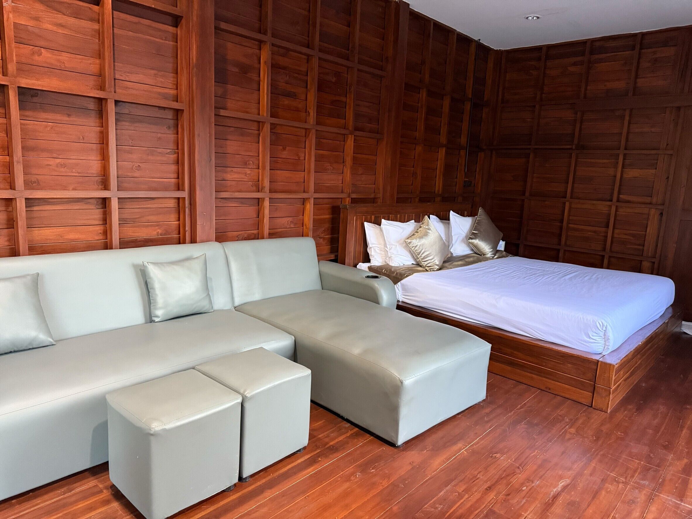 A modern bedroom with a wooden panel wall, a grey sectional sofa, a single bed with white bedding, and a wooden floor.