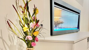 Un jarrón de mimbre con flores sobre una consola blanca, junto a un televisor de pantalla plana sobre una pared blanca.