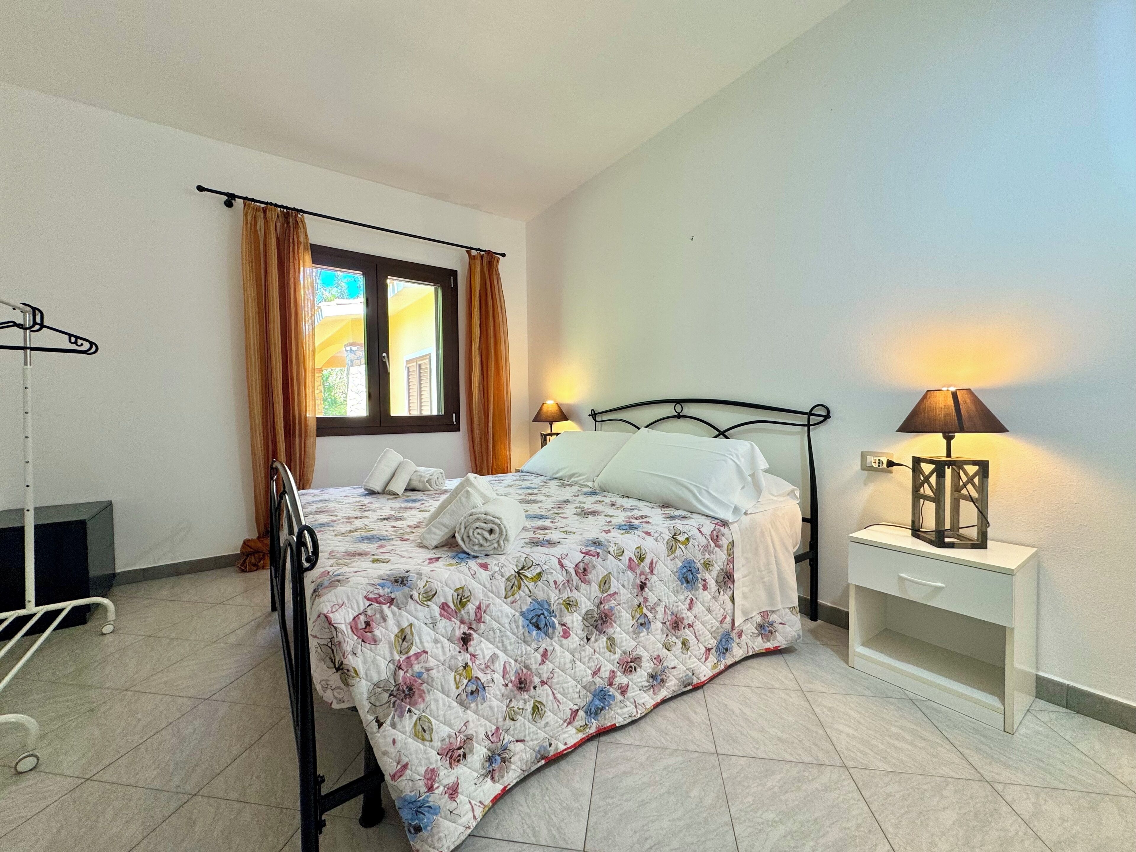 Un lit bien fait, recouvert d’une courtepointe à motifs floraux, une lampe de chevet et une fenêtre avec des rideaux.