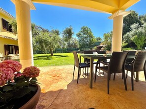 An outdoor patio with a table and chairs, surrounded by yellow columns, and a view of a green lawn and trees. - " Villa with pool Luxury " (Cardedu)