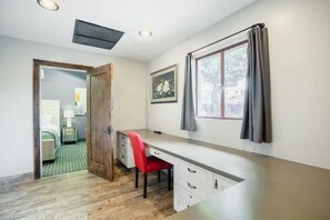 Une pièce comprenant un bureau, une chaise, une fenêtre avec des rideaux, une porte donnant sur une autre pièce et un tableau encadré accroché au mur.