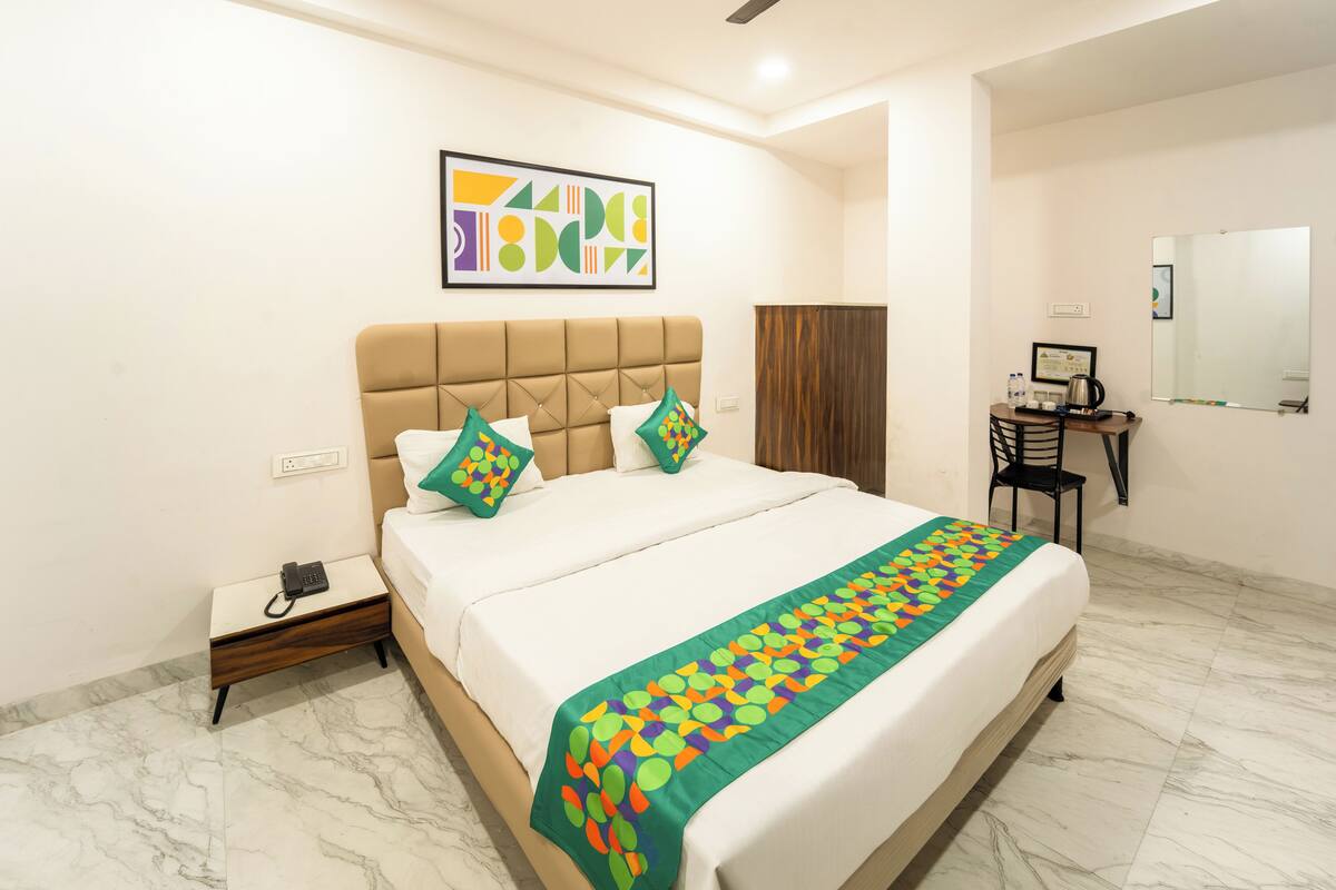 A hotel room with a bed, a desk, a chair, a ceiling fan, a mirror, and a painting on the wall.