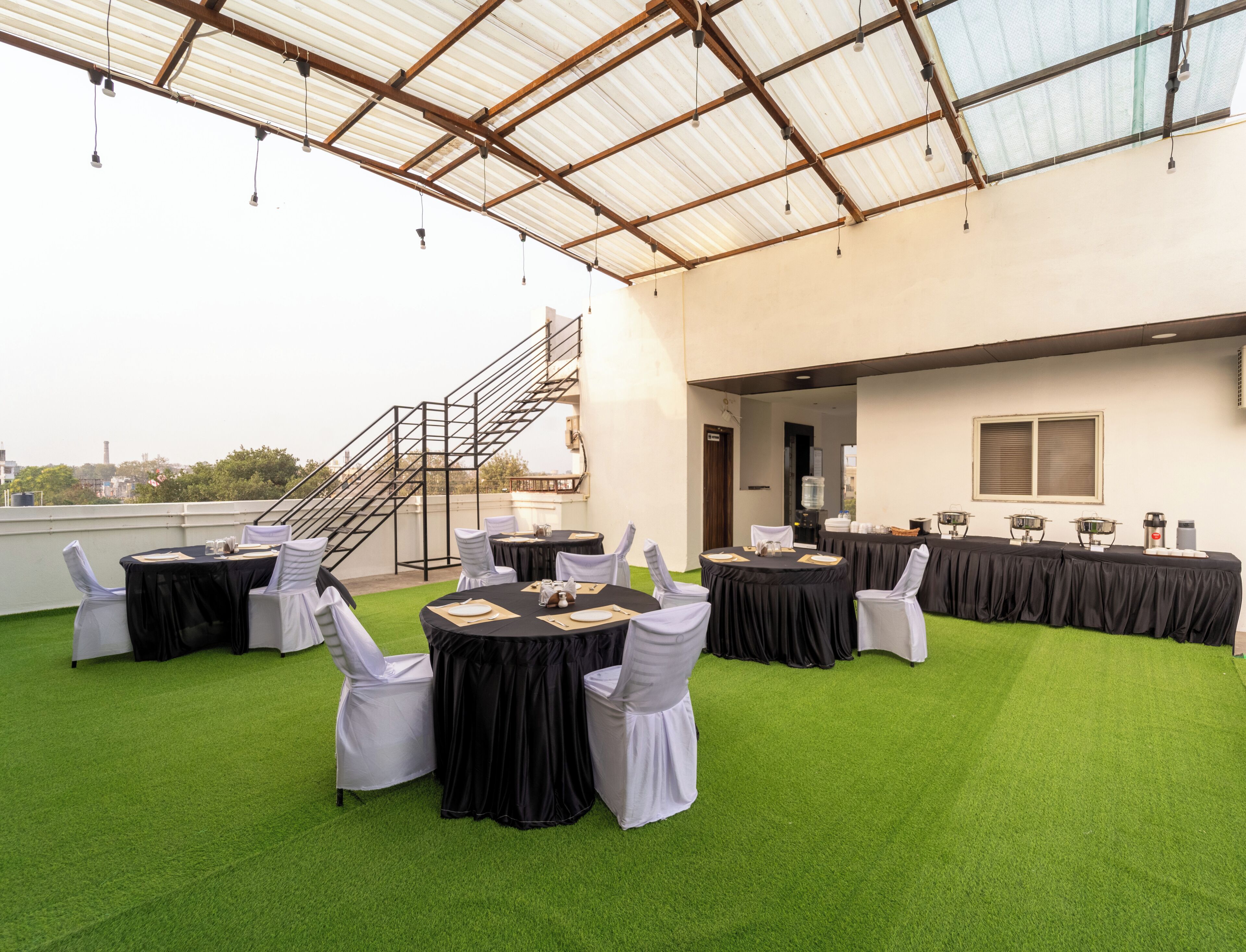 A rooftop event space with multiple round tables, white chairs, and a staircase leading to an upper level.