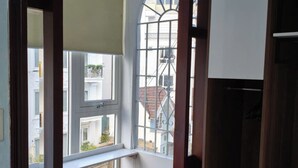 Une pièce avec un plancher en bois, une fenêtre donnant sur des immeubles, une chaise et une armoire en bois.