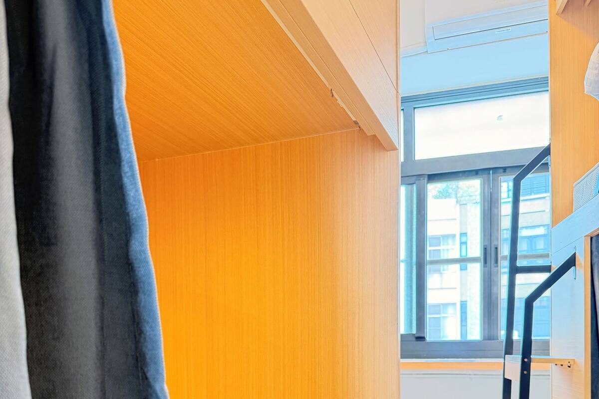 Ein Etagenbett mit Holz Kopfteil, ein Fenster mit Blick auf Gebäude und eine schwarze Metallleiter.