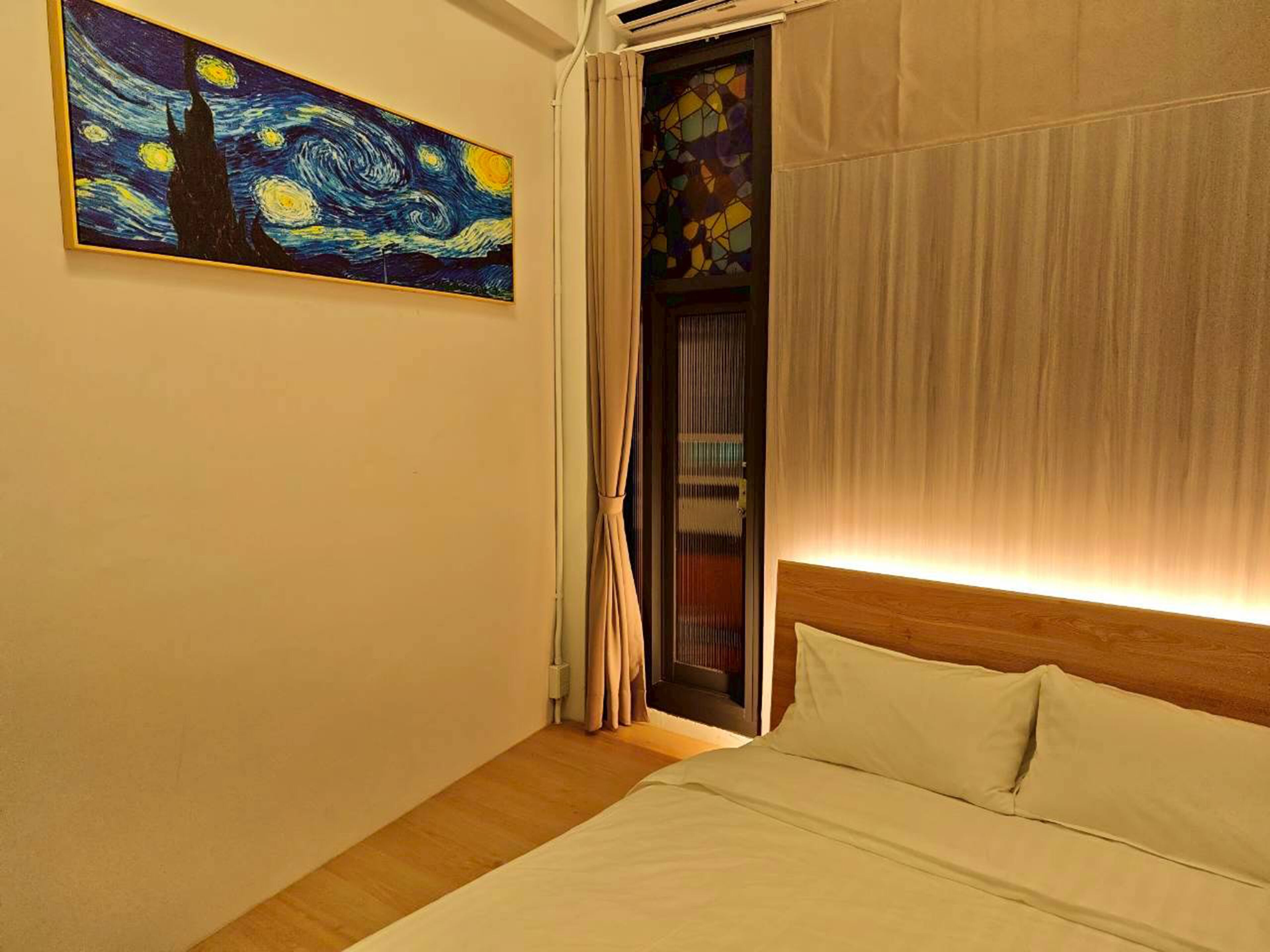 A hotel room with a bed, a painting on the wall, a window with curtains, and a wooden headboard.
